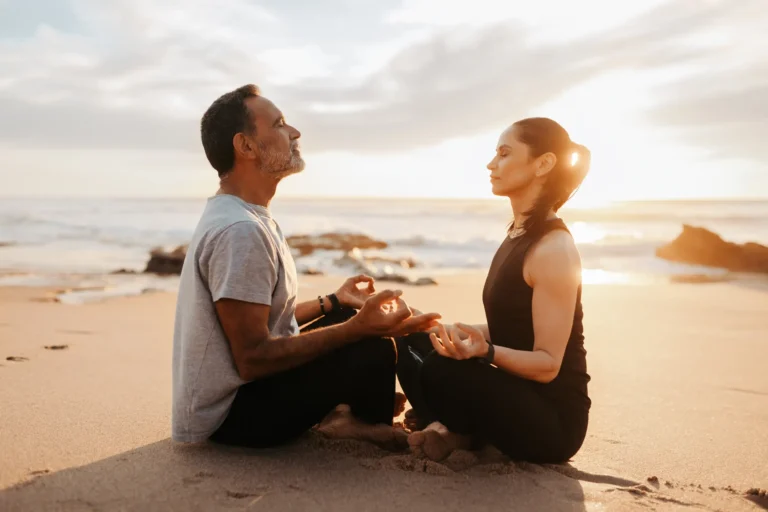 A man and a woman meditating near the seashore on the beach.